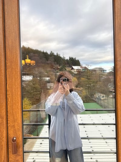 An individual, identified as an adult female, is captured in a self-portrait reflection within a large window or glass door. She is positioned centrally, holding a black-cased smartphone with a visible multi-lens camera array with both hands, pointing it towards the reflective surface to photograph the scene. Her face is largely obscured by the phone and dark hair. She wears a light blue, collared, button-up shirt over a lighter undershirt and light-wash denim trousers.

The reflection's interior frame reveals sections of a light-brown wooden window or door frame with vertical grooves, a brown handle on the left, and part of a golden-colored three-shade chandelier in the upper-left.

The exterior view, consistent with Bakuriani, Georgia, presents a landscape under an overcast, grey sky. Hilly terrain, densely covered with a mix of deciduous trees (some with yellow foliage, others bare) and evergreen conifers, rises in the background. Scattered patches of light snow are visible on the ground and rooftops. Several buildings are integrated into the slopes, including a structure with a light-colored roof and another with a multi-paneled, discolored roof. In the immediate foreground of the exterior scene, a black metal balcony railing is visible, beyond which lies a bright green artificial turf area, marked with white lines, indicative of a sports court (likely tennis). Further structures are visible beyond this court.