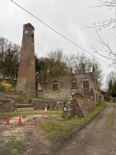 This image depicts the ruins of former industrial buildings at Snailbeach Lead Mine, Snailbeach, United Kingdom, under an overcast sky. The central subject is a tall, reddish-brown brick chimney with a stone base, standing to the left of a larger, partially collapsed stone and brick building. This building features multiple arched openings, indicating former windows or doorways, and its roof is absent. Further remnants of stone and brick walls and foundations are present in the foreground and to the right of the main ruin.
No individuals are visible. In the foreground, on a muddy and grassy patch, there are three traffic cones: two small, solid orange cones and one taller, red and white striped cone positioned behind them, secured by a white rope or tape. A wooden post with a small sign attached is visible near the taller cone, but its specific text or imagery is indistinguishable.
A narrow, unpaved dirt path extends from the foreground along the right side of the buildings, curving slightly to the right and disappearing into the distance. Bare deciduous trees cover the steep hillsides behind the ruins, and a single black cable or wire stretches diagonally across the upper portion of the frame. A patch of yellow flowering shrubs grows near the base of the chimney. In the far left background, a white building with an outdoor deck and furniture is visible on the hillside.