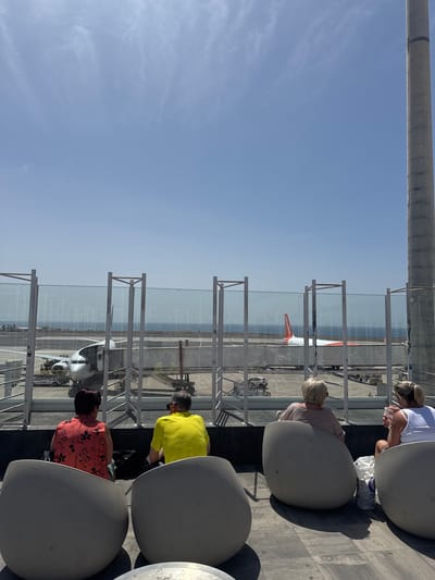 Four people sit in chairs on a rooftop overlooking an airport in San Bartolomé, Spain. A white metal fence with glass panels lines the rooftop. An airplane is visible on the tarmac. The sky is blue. A thick, gray support beam runs along the top of the image.