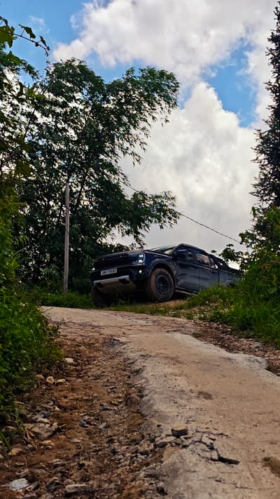 The image captures a black Ford Ranger pickup truck ascending a rugged dirt road in Hoàng Su Phì Commune, Vietnam. The truck is angled upwards, its front end prominently visible, with the license plate "29K-179.83" displayed. The tires are caked with mud, indicating recent off-road travel. The road surface is uneven, a mix of packed earth and exposed rocks. Lush greenery borders the road on both sides, with trees partially obscuring the truck. A utility pole stands to the left of the truck. The sky overhead is partially cloudy, with patches of blue visible through the breaks.