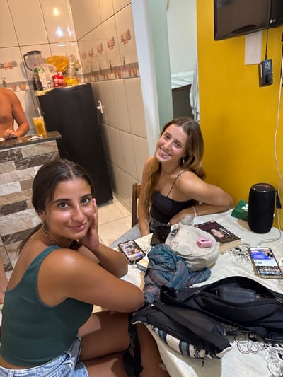 Two young women pose for a selfie. One woman wears a black tank top and has long brown hair. The other is closer to the camera and has darker hair and a green tank top. They sit at a table covered with items including a phone, book, speaker, and bag. A kitchen is visible in the background with a countertop, tiles, and food items. A shirtless person stands by the counter. The photograph is slightly tilted.