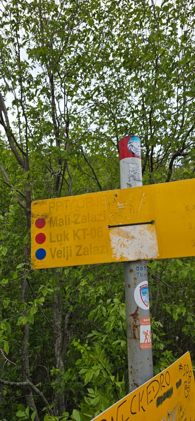 Here is a description of the image:
The image shows a trail marker, likely in a wooded area within the vicinity of Kopito, Montenegro. The trail marker, bright yellow in color, is attached to a metal pole. The marker displays information about trails: "PPT: ORJEN Mali Zalazi Luk KT-06 Velji Zalazi". Three colored dots, two red and one blue, are visible at the bottom of the sign. Stickers and signs are affixed to the metal pole. The background is predominantly composed of green foliage, suggesting a wooded environment.
