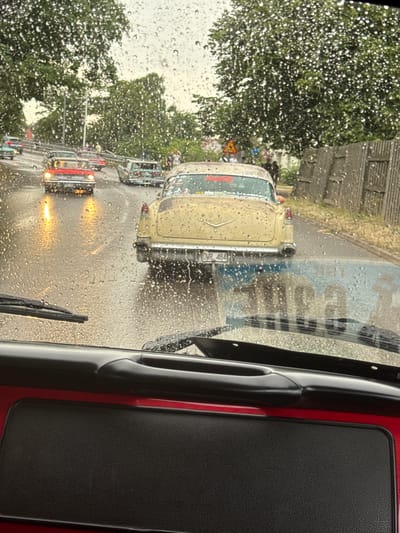 The image captures a view from inside a vehicle looking out through a rain-streaked windshield. It appears to be a procession or gathering of vintage cars on a wet street. The dominant subject is a yellow vintage car in the foreground, viewed from the rear, with the license plate "DRK 484" visible. To its left, a red vintage car is visible with its headlights on, reflecting on the wet asphalt. Further down the road, other vintage cars are present, and several people are standing on the roadside. The environment suggests an overcast day with rain, indicated by the water droplets on the windshield and the wet road surface. The presence of trees lining the street and a wooden fence on the right side of the road suggest an urban or suburban setting. Given the context of a vintage car event and the specific license plate, this scene is likely taking place in Falkenberg, Sweden, a location known for such gatherings.