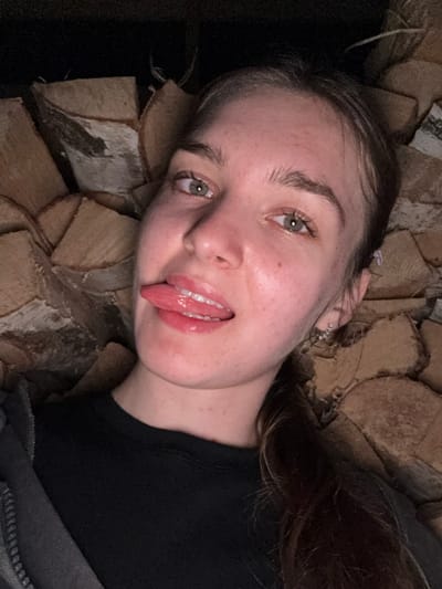 A young woman with light brown hair and green eyes is shown in a close-up, horizontal, selfie-style shot. She is wearing a black t-shirt and a gray hooded jacket with a zipper. Her tongue is sticking out and she is wearing lip gloss. She has a small, sparkling earring in her left ear. She is leaning against a wall of stacked logs, which appears to be outdoors. The lighting is dim, suggesting it may be nighttime or an indoor setting with minimal light. The background of stacked logs is textured and varies in color from light brown to darker brown, with some showing signs of bark and wood grain. The image does not provide any specific clues to its location in the city of Nizhniye Muly, Russia.