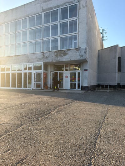 The image shows the exterior of a building in Pyt'-Yakh, Russia. The building is made of concrete and has large windows. A person is walking towards the entrance of the building, which has glass doors. The ground in front of the building is asphalt. The sky is clear and blue. There are some metal bars and a ladder attached to the side of the building. The overall impression is of a utilitarian building, possibly an industrial facility or an office building. The sunlight suggests it is daytime.
