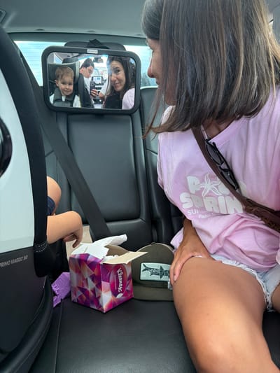A woman in a pink t-shirt and denim shorts sits in the passenger seat of a car, turned to her right. A car seat mirror is attached to the headrest behind her, reflecting a young boy in a car seat, a man taking a photo with his phone, and the woman’s own reflection. A box of Kleenex tissues and a green baseball cap are on the seat next to her. The image appears to have been taken inside a vehicle, possibly in the city of El Prat De Llobregat, Spain, as indicated by the context.