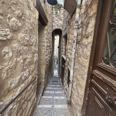 This image captures a narrow, stone alleyway in Saint-Paul-de-Vence, France. The perspective is looking upwards and into the alley, with the rough, textured stone walls dominating the foreground and sides. A white pipe runs horizontally along the top of the alley, adorned with numerous stickers of varying designs and colors. Some of these stickers appear to be logos or brand names, while others are more artistic or humorous.

The alley itself curves slightly as it recedes into the distance. On the right side, a set of stone stairs with pebble inlays leads upwards, creating a sense of depth and perspective. The architecture suggests an old European town, with weathered wooden elements visible at the very top of the frame, possibly part of a door or window.

In the background, the alley opens slightly, revealing more stone buildings and glimpses of greenery, including some plants with dark red foliage. There's a hint of sunlight, suggesting it might be daytime, though the narrowness of the alley creates significant shadow. The overall impression is one of an ancient, well-trodden path, with the stickers adding a touch of modern street art to the historical setting. No people are visible, and the scene conveys a sense of quiet exploration and discovery.