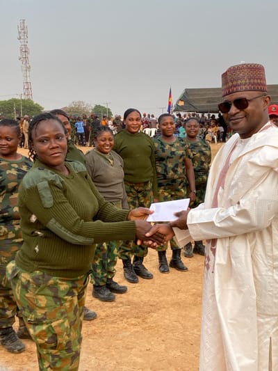 A line of women in camouflage uniforms and black boots stand facing right. A man in a traditional white robe and patterned cap gives one of the women a white envelope. The woman accepting the envelope is smiling. There is a crowd of people in the background. In the bottom left of the frame is a tall metal structure.
