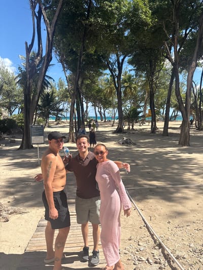 The scene depicts three individuals posing on a wooden boardwalk at a beach in Wellhouse, Barbados, under a clear blue sky with scattered clouds.
From left to right:
1. A male individual, shirtless, wearing black shorts and a black baseball cap. Tattoos are visible on his left bicep and right lower leg. His right arm is around the waist of the central individual.
2. A male individual wearing a brown polo shirt and khaki shorts. He holds a clear plastic water bottle in his left hand and has an arm around the waist of each person beside him.
3. A female individual wearing a long, light pink, textured long-sleeved dress and sunglasses. Her left arm is around the waist of the central individual.
All three individuals are smiling and looking towards the camera. The wooden boardwalk extends from the bottom of the frame into the mid-ground. The surrounding ground is sandy. Numerous tall trees, including palm trees and other broadleaf species, are dispersed across the sandy area, casting dappled shadows. A white sign labeled "BEACH RULES" on a post is visible to the left of the central individual. In the background, beyond the trees and sand, the turquoise ocean is visible, with a few other individuals walking on the beach. A yellow structure, possibly a lifeguard stand, is visible on the distant sandy beach to the right.