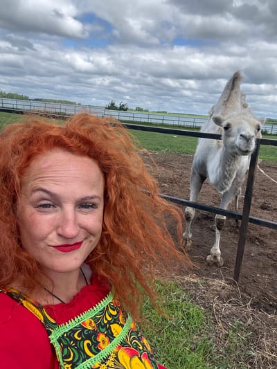 A fair-skinned woman with bright red, curly hair and red lipstick is taking a selfie with a camel. She is wearing a red top with a vibrantly colored, embroidered apron. The camel, which is pale gray, stands behind a metal fence in a muddy enclosure. The background shows a cloudy sky, a body of water, a fence, and some trees. The scene appears to be outdoors, possibly at a zoo or animal sanctuary, in or near the city of Tri Ozera, Russia.