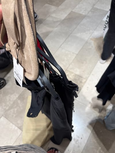 The image shows a close-up of clothing items hanging on hangers in what appears to be a retail environment. In the foreground, a person is holding up a tan-colored garment, possibly made of suede, with a white tag attached. Behind this item, black garments are also hanging on hangers, one of which appears to have a tear. The background is blurred but shows a tiled floor and hints of other people and clothing in what resembles a department store or clothing boutique. The lighting suggests an indoor setting. There is no information in the image to determine the location as Dartford, United Kingdom.