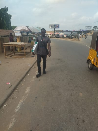 The image shows a man walking on a paved road in Maraba, Nigeria. He is wearing a dark gray t-shirt and black jeans, carrying a large green and white object, presumably a folder or book, under his left arm. He also has a black satchel slung over his left shoulder and a watch on his left wrist. The road is lined with a brown dirt shoulder, separated by a curb. On the left side of the image, there are several tables and stalls with people selling various items. There are also several other people walking on the road in the distance, including one person riding a motorcycle. The image shows a yellow tricycle, with a yellow and red stripe, stopped on the right side of the road. The sky is overcast with a light gray sky, and there is a tall billboard in the background. The scene suggests an urban environment with the presence of stalls and traffic, including a tricycle.  The image was taken on a slightly cloudy day, and the overall atmosphere is one of daily life.