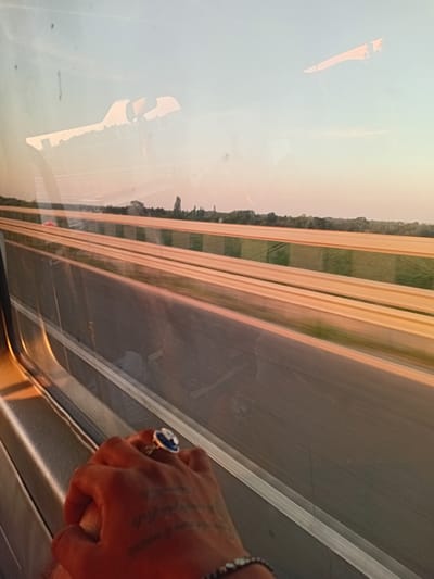 The image depicts a view from inside a vehicle, looking out a window. The hand of a person, adorned with a ring featuring a blue stone, is visible in the foreground. In the background, a blurred landscape of fields and trees is seen, suggesting movement. The sky is a gradient of light blue and pink, indicating either sunrise or sunset. There is a reflection in the window, showing the interior of the vehicle, including what appears to be a car's dashboard and side mirror. The location is identified as Hegyeshalom, Hungary, which is known for its border crossing with Austria and its role as a transportation hub. The blurriness of the background implies the vehicle is in motion.