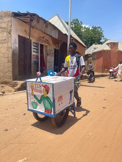 In Magajin Gari A, Nigeria, a young man, likely an ice cream vendor, is pushing a handcart decorated with colorful drawings of ice cream and an apple. He wears a white jersey with black and yellow accents, featuring the "Emirates Fly Better" logo and "R" logo. The cart has two large wheels and a white box adorned with a drawing of a person holding an ice cream cone. A blue megaphone rests on top of the cart. The scene takes place on a dusty dirt road lined with modest buildings. One building, with a damaged corrugated metal roof and stone walls, has signs with text including "BABAN K. JAMALU" and "PATENT MEDICINE STORE". In the background, other people are visible, along with parked motorcycles. The sky is bright and blue with scattered clouds.