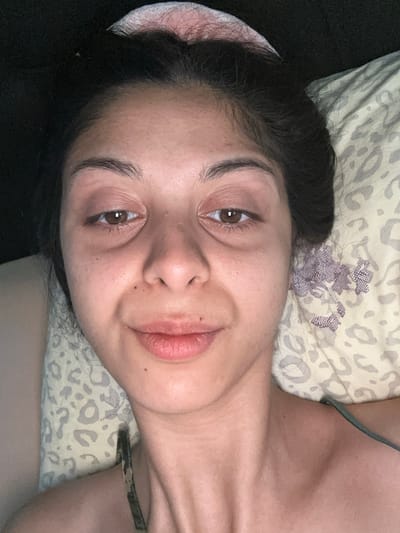 The image is a close-up, high-angle shot of a young woman lying on a pillow. She has dark hair pulled back and is wearing a light-colored garment. Her eyes are open and looking at the camera. The background consists of a black surface and a pillow with a light-colored, patterned fabric, possibly leopard print and floral motifs. The woman's face is illuminated, suggesting an indoor setting, possibly a bedroom. There are no clear indicators of the city of Augsburg, Germany, within the image itself.