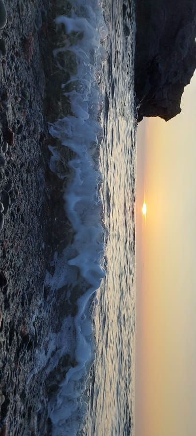 This is a vertical, close-up, outdoor shot of a rocky shoreline at sunset. The left side of the image features dark, wet rocks of various sizes and textures, with a wave breaking over them and creating white foam. The water is dark and appears choppy. To the right of the water, the sky transitions from a pale yellow near the horizon to a soft orange, with the sun visible as a bright, small circle low on the horizon. A large, dark rock formation juts into the frame from the upper right corner. The scene is devoid of any people or animals. The overall impression is one of natural beauty and tranquility at the end of the day, with the elements of water, rock, and sky in focus.