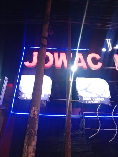 A nighttime shot reveals a lit sign for "JDWAC JR." vertically aligned. Below the name, two smaller signs are visible, one labeled "PORK CHOMA & KACHUMBARI" and the other "KUKU & KACH." The signs are illuminated against a dark backdrop with blue neon lighting that adds contrast. There are poles and wires that obscure the signage to a degree.