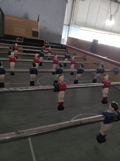 This is a low-angle, medium shot of a foosball table. The table is green with white lines, and the figures are red and blue. The foosball players are arranged in rows on metal rods. In the background, there is a cardboard box with "LG Microwave Oven" printed on it, and a fluorescent light fixture. The scene appears to be indoors in a dimly lit space. The image is of a foosball table located in Chaponost, France.
