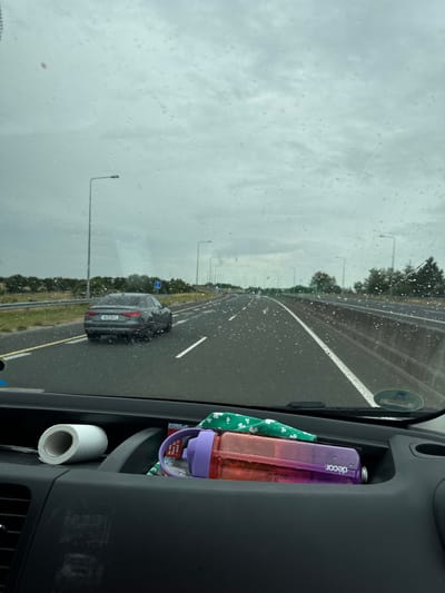 The image depicts a view from inside a vehicle looking forward through a rain-streaked windshield onto a highway. A dark-colored Audi sedan is visible driving in the right lane ahead. The highway has multiple lanes and is lined with trees and streetlights. In the foreground, inside the vehicle, a roll of toilet paper and a reusable water bottle with a purple lid and orange body are visible in a compartment. The sky is overcast and grey. The location is identified as Dunbell, Ireland.