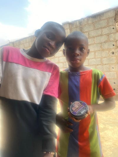 Two young individuals, likely children, stand outdoors in Idiroko, Nigeria, facing the camera. The individual on the left wears a long-sleeved color-block shirt in pink, white, and black, with a light-colored beaded bracelet on the left wrist. The individual on the right wears a vertically striped short-sleeved shirt in red, blue, purple, green, and yellow, holding a black circular container labeled "KAIMA HAIR WAX REGULAR BLACK" in their left hand, displaying the label. Both individuals have closely cropped dark hair and look directly forward. The background consists of a light-colored concrete block wall behind them, with a partially cloudy sky visible above the wall. The ground appears to be a light-colored, flat surface, suggesting packed earth or concrete.
