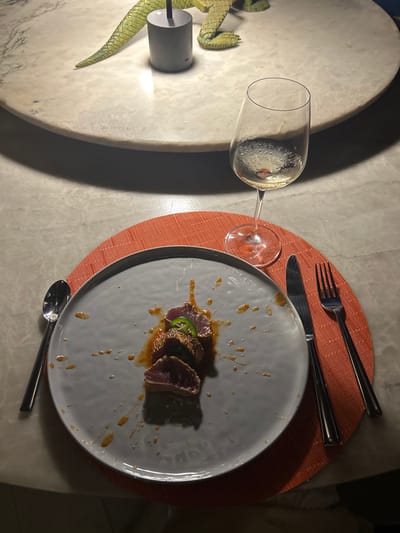 The image shows a plate of food on a table at a restaurant in Playa Herradura, Costa Rica. The plate contains three slices of seared tuna, topped with a green jalapeno pepper and sesame seeds. The tuna is arranged vertically, with the jalapeno pepper resting on the top slice. The plate is surrounded by an orange placemat. A glass of white wine is sitting on the table next to the plate. To the left of the plate is a silver spoon and to the right is a silver knife and fork. There is a white tablecloth and a decorative, ceramic crocodile on the table, which sits in front of a grey lamp base. The scene is lit from above. The image was likely taken during the evening. The location in the city is an outdoor dining area at a restaurant or bar.