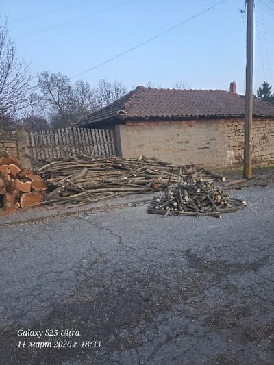 The image depicts an outdoor, static scene in Dolna Beshovitsa, Bulgaria, captured on March 11, 2026, at 18:33 by a Galaxy S23 Ultra device.
The foreground consists of a cracked and worn asphalt road surface, sparsely scattered with wood chips and sawdust. Two accumulations of cut wood are present on this surface. To the left, a larger, elongated pile of diverse branches and logs extends horizontally. To the right, a smaller, conical pile of shorter, more uniformly cut wood pieces is situated. A small, two-wheeled handcart is partially visible, resting within the larger wood pile.
In the midground, a weathered wooden picket fence stands between the road and a single-story building. The building features a facade of mixed construction, appearing to combine sections of brickwork with areas of what resembles rendered or mud-brick material. It is topped with a sloped roof composed of terracotta tiles, and a brick chimney stack is visible on the roofline towards the right. Bare deciduous trees are positioned behind the fence and the building, indicating a late winter or early spring environment.
The background shows a clear, pale blue sky with faint overhead power lines traversing the frame. A tall, dark wooden utility pole is prominently situated on the right side of the image, extending upwards beyond the roofline of the building. A coniferous tree is partially visible on the far right edge of the frame. No individuals or active interactions are present in the scene.