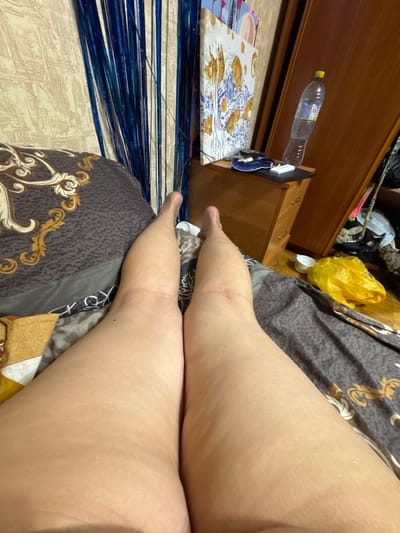 The image, taken in Tarasovka, Russia, shows a first-person perspective of a person's legs resting on a bed. The legs are positioned in the center of the frame, with the right leg slightly closer to the viewer. The bedspread has a dark brown and gold ornate pattern. To the left of the bed, a wall with a textured beige wallpaper is visible, adorned with vertical blue tinsel. Behind the tinsel, a framed artwork with yellow and blue elements is partially visible. To the right of the bed, a wooden dresser with drawers stands against a wooden wall. On top of the dresser, a plastic bottle of water and a small white electronic device are present. Further to the right, a yellow plastic bag and a white cup are on the floor, near a closet filled with clothes. The lighting suggests an indoor environment, possibly a bedroom.