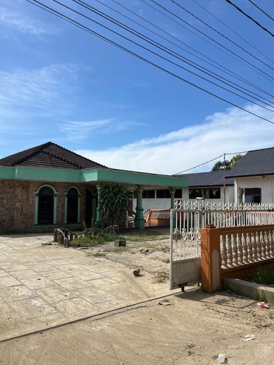 The image shows a side view of two houses next to each other, with a clear blue sky and some wispy clouds in the background. Multiple electrical wires run diagonally across the sky.

The house on the left has a dark tiled roof and a light green trim. Its walls are made of stone-like material, and it features two arched windows with dark panes. The house on the right has a metal roof and a white exterior. It has a white balustrade with decorative elements and a brown pillar. Part of the house's interior is visible through the windows, showing what appear to be framed pictures of people. A plant is visible in front of the right house.

The ground in front of the houses is a mixture of paved concrete tiles and dirt. Tire tracks are visible in the dirt. There is some scattered debris on the ground, including a white plastic cup.

The scene appears to be outdoors, likely in a residential area. The lighting suggests it is daytime, and the clear sky indicates fair weather. The houses and the style of the balustrade suggest a location in Indonesia, as indicated by the provided context. There is no visible text in the image.