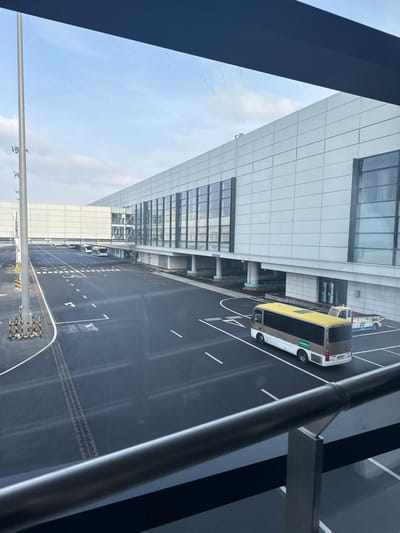 The image displays an exterior view of the tarmac and terminal building at Ürümqi Diwopu International Airport (URC) in Ürümqi, China, observed from an elevated vantage point through a window. A dark metal railing is visible in the immediate foreground, spanning the lower portion of the frame.

The environment consists of a partially cloudy sky and a dark asphalt tarmac appearing damp, marked with distinct white dashed lines, solid lines, arrows, and the letter "A". On the left, a tall, light-colored utility pole with yellow and black striped barriers at its base stands beside the tarmac.

The background is dominated by a modern airport terminal building featuring light-colored exterior panels and numerous dark, rectangular glass windows. An elevated, enclosed walkway or sky bridge, also light-colored, extends from the main terminal structure towards the left.

Multiple vehicles are present. In the mid-ground on the right, a two-tone (brown/dark grey bottom, yellow top) airport shuttle bus with dark windows is positioned on the tarmac, facing away from the viewer. Immediately to its right, a white ground support equipment vehicle is partially visible. Further left, beneath the elevated walkway, two additional light-colored airport buses are parked. No individuals are discernible in the scene. The overall atmosphere suggests an operational but uncrowded airport setting.