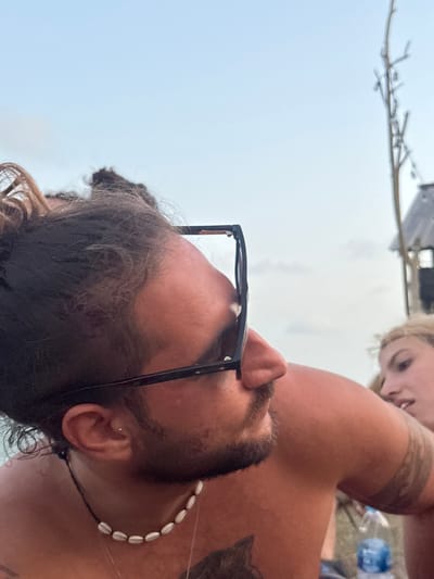 The image captures a close-up, profile view of a male individual positioned in the foreground, outdoors in Ban Pang Pring, Thailand. The male has dark hair tied back in a bun, a beard, and wears black square-framed sunglasses pushed onto his forehead. A white seashell necklace is visible around his neck, and a silver stud earring is present in his left ear. He bears a dark tattoo on his upper left chest and a tribal-style tattoo on his right upper arm. To his right and slightly behind him, a blonde-haired female is partially visible, looking towards the male. Below her right arm, a clear plastic water bottle with a blue cap is present. The background shows a light blue sky with scattered faint clouds. On the far right, a tall, slender wooden structure with branches extends upwards, and a dark, partially visible structure, possibly a hut, is situated below it.