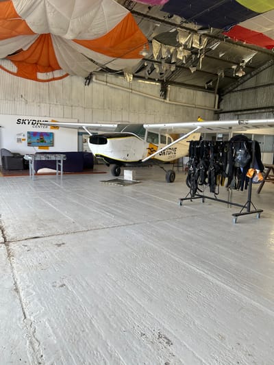 The image displays the interior of a skydiving facility located in Chascomús, Argentina. The foreground consists of a light grey concrete floor exhibiting wear and visible seams. In the midground, a single-engine propeller aircraft, predominantly white with yellow accents and "SKYDIVE CENTER" branding on its fuselage, is parked. The aircraft's nose cowling is detached, exposing internal engine components. It is positioned on a grey block beneath its front landing gear and a metal plate under its rear wheel. To the left of the aircraft, a lounge area is arranged against a white wall, featuring a dark grey armchair, a dark blue couch, and a low, light-colored table. A television screen on the wall displays an animated cartoon image, and a "SKYDIVE CENTER" logo with a QR code is also visible. To the right of the aircraft, a mobile, black metal rack holds multiple black skydiving harness rigs. The background reveals corrugated metal walls and a high ceiling structure. Numerous large, colorful parachutes (including orange, white, blue, green, yellow, and red) are partially unfurled or draped from the ceiling, alongside decorative or insulating foil-like materials and exposed industrial light fixtures. No individuals are present within the scene.
