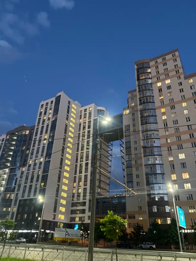 The image captures a view of modern high-rise apartment buildings in Dachnoe, Russia, during twilight. The sky is a deep blue with scattered clouds. The buildings are tall and feature a mix of light-colored facades and extensive glass windows, many of which are illuminated from within. A prominent architectural feature is a connecting bridge-like structure between two of the buildings. In the foreground, a street with cars is visible, separated from the viewpoint by a metal fence and a grassy area. Streetlights and power lines are also present in the scene. Signs in Russian are visible on the side of the road. The overall impression is of a residential urban environment at dusk.