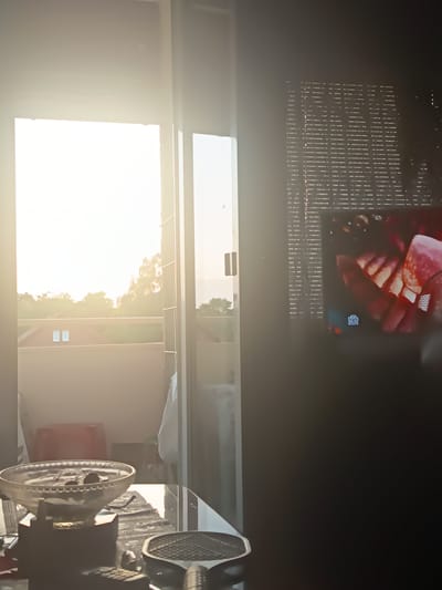 The image depicts a domestic indoor scene, likely an apartment or house, with a view of a balcony or exterior through a large glass door or window. The time of day appears to be late afternoon or early evening, with bright sunlight streaming in from the left, casting a warm glow on the interior and silhouetting some objects.
On the left side of the foreground, a glass bowl filled with dark, circular objects, possibly cherries or olives, sits on a glass-topped table. A dark, silhouetted object, possibly a small appliance or decorative stand, supports the bowl. To the right of the bowl, a portion of a patterned mat is visible on the table. Also on the table, a black object resembling a small paddle or a racket is partially visible, along with what appears to be a remote control. A red, rectangular object is visible in the mid-ground on the left, possibly a piece of furniture or a storage container.
Through the glass door, a balcony railing is visible. Beyond the railing, there is a view of trees and buildings, suggesting an urban or suburban setting. Two small, rectangular windows are visible on a building in the mid-ground outside.
On the right side of the image, a television screen is on and displays a close-up image of what appears to be food, possibly strawberries being prepared. The television is mounted on or placed against a dark wall, with horizontal blinds or a textured surface behind it. A faint logo, appearing as Cyrillic characters, is visible on the television screen, suggesting it might be a Russian-language channel.
The overall impression is of a lived-in space, with a relaxed atmosphere, possibly during a time of leisure. There are no people clearly visible in the image. The lighting suggests a sunny day, and the presence of a television indicates modern living. The image does not provide any specific landmarks or identifying features that would definitively place it in Oldenburg in Holstein, Germany, beyond the possibility suggested by the user's prompt.