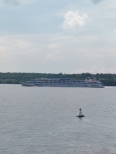 A large white cruise ship with multiple decks and blue trim is sailing on a wide, calm river. The ship has many windows and several lifeboats visible on its upper decks. The river is choppy with sunlight reflecting off its surface. In the foreground, a white buoy with a black top floats in the water. In the background, a dense line of green trees forms the shoreline. The sky above is cloudy with patches of blue visible. This scene is likely located on the Volga River in Uglich, Russia, given the context of the provided image.