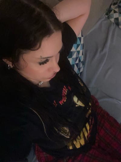 The image displays an individual lying prone on an upholstered surface within an indoor setting in Saint-Leu-la-Forêt, France. The person has long, dark hair, parted on the side, extending over the shoulders. Facial features visible in profile include a fair complexion, a prominent nose with a silver piercing in the left nostril, and full lips. The left eyebrow features a piercing with two small silver balls. Long, dark eyelashes are apparent on the left eye. The left ear holds multiple silver hoop earrings. Around the neck, a pearl necklace is present. The individual wears a black t-shirt with a graphic print featuring red/orange text, partially legible as "WOLVES!", above a stylized face and further yellow text. Red and black plaid pants or shorts are visible from the waist down. The left arm is bent, with the hand positioned behind the head or neck. The individual rests against a light grey or blueish-grey upholstered surface. A pillow with a geometric pattern of blue, grey, and white triangles is visible behind the head. Another solid grey section of upholstery is to the right. The scene is dimly lit.