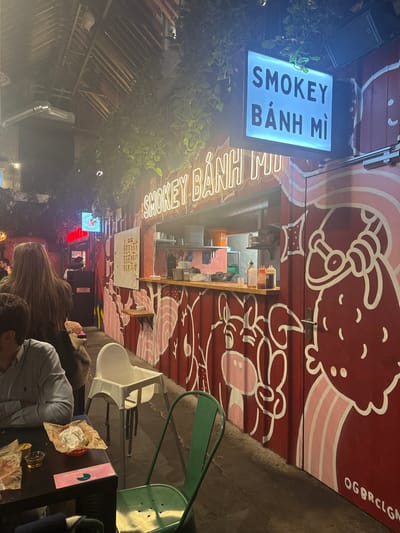 The image depicts an indoor food stall, "Smokey Bánh Mì," located in what appears to be a covered food market or hall in Saint-André-lez-Lille, France. The stall is constructed from dark red corrugated metal panels, adorned with stylized white and pink cartoon illustrations of pigs or cows. A prominent neon-style sign above the counter reads "SMOKEY BÁNH MÌ," supplemented by a perpendicular illuminated sign displaying the same text. The counter features various condiment bottles and food preparation items. To the left of the stall, two individuals are seated at a dark rectangular table. One person, with long light brown hair, is seen from the back, while another is partially visible from the side, wearing a striped shirt. On their table rests a red basket containing a wrapped food item and a clear plastic cup. A white plastic high chair is positioned between the dining table and the food stall, and a dark green metal chair is in the immediate foreground. The environment includes exposed wooden ceiling beams, ductwork, and hanging green foliage. The scene is illuminated by artificial lighting, suggesting an evening setting. The letters "OGBRCLGN" are visible in white paint on the lower right door of the food stall.