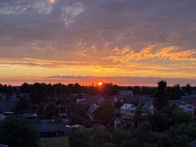 The image depicts a sunset over a village in the Pratasevichy Village Council, Belarus. The sky is filled with colorful clouds, ranging from deep purple to vibrant orange and yellow, as the sun is low on the horizon, casting a warm glow. Below the sky, a dense line of trees forms the edge of a settlement. The village consists of numerous houses with pitched roofs, some appearing older with corrugated metal roofing, while others are more modern. The architecture suggests a rural or suburban setting. The foreground is dominated by dark green foliage, indicating a dense growth of trees and bushes, possibly within gardens or natural areas surrounding the houses. The overall scene is tranquil, characteristic of a rural evening. There are no people visible in the image.