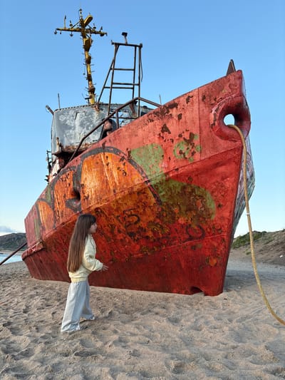 In Teriberka, Russia, a large, rusted red ship rests on a sandy beach. A young girl with long, light-brown hair stands facing the ship, wearing a pale yellow jacket and grey sweatpants. Her right hand is extended towards the vessel. Behind her and to the left, the water is visible, meeting a rocky, treeless coastline. A thick, yellow rope is tied to the ship's bow and trails onto the sand. In the upper section of the ship, a person wearing a black hooded sweatshirt looks upwards. The sky is clear and blue. The ship is covered in graffiti and shows significant wear and tear.
