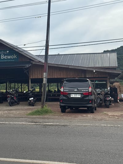 The image depicts a roadside scene in Lampisang, Indonesia, likely in front of a restaurant or food stall. A black Toyota Avanza with license plate BL 1690 AD is parked in the foreground. Behind the car, a rustic, open-sided building with a corrugated metal roof is visible. Several motorcycles are parked in front of the building, suggesting customers. The overall environment appears to be rural or semi-rural, with a dirt or gravel lot surrounding the building and a paved road in the foreground. A utility pole stands near the building. In the background, a tree-covered hill is visible. The sky is overcast.