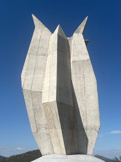 The image shows a concrete monument or sculpture in Godinje, Montenegro. The sculpture is composed of stacked, angular concrete segments, forming a sharp, geometric shape against a clear blue sky. The concrete surface shows signs of weathering and exposed aggregate. Part of the monument abuts a whitewashed element that includes vegetation from surrounding hills on the right side of the frame.