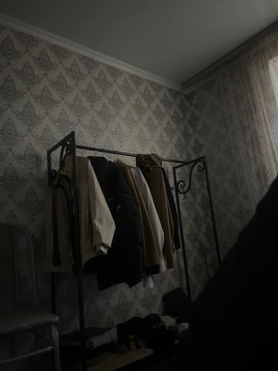 A dimly lit room features a black metal garment rack filled with various jackets and coats. The walls are covered in a gray and white damask wallpaper pattern. To the left, a portion of a chair with a patterned seat is visible. On the floor beneath the garment rack, several items are present, including folded clothing and containers. A sheer curtain hangs on the right side of the image, covering a window. The scene suggests a domestic setting within a room in Первомайское сельское поселение, Ukraine.