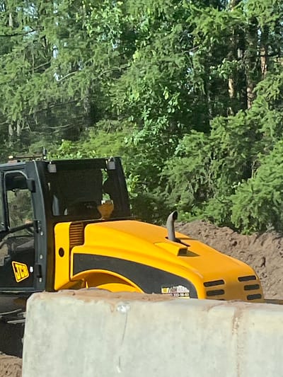A yellow and black JCB construction vehicle is positioned near a textured white wall, with trees filling the background. The location is Staryi Chul'tem, Russia.
