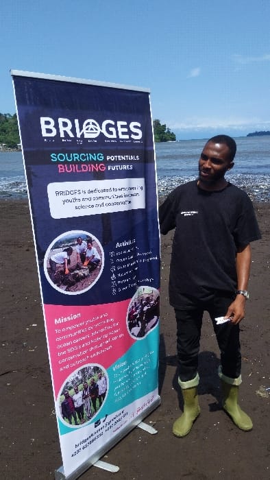 A man in a black t-shirt, black pants, and green boots stands to the right of a banner that reads "BRIDGES: SOURCING POTENTIALS BUILDING FUTURES". The banner is situated on a dark sandy beach with a body of water in the background. The man is holding a white card in his right hand. The banner includes a mission and vision statement, as well as pictures of people participating in activities. The banner also lists the following activities: "Social Action", "Environmental Awareness", "Youth Parliament", "Volunteerism", "Marine Research", "Nature Tours", "5km Run". The location is Limbé I, Cameroon at coordinates 4.004 latitude and 9.211 longitude.