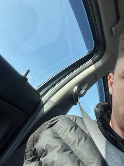 The image displays the interior of a vehicle under bright, clear daylight conditions. The right side of a male individual's head and upper torso are visible in the foreground, wearing a black puffer jacket and a properly fastened grey seatbelt across the chest. Overhead, a large panoramic sunroof is open, revealing a clear, bright blue sky. The vehicle's interior is detailed with a black textured car seat in the lower left, a black door panel on the far left with a visible black door lock knob, and grey interior trim components, including the A-pillar and roofline. Direct sunlight illuminates the scene. No specific landmarks or environmental identifiers are present in the image to determine a precise location within the city of Opalchenets, Bulgaria; the scene is confined to the vehicle's interior within the general area.