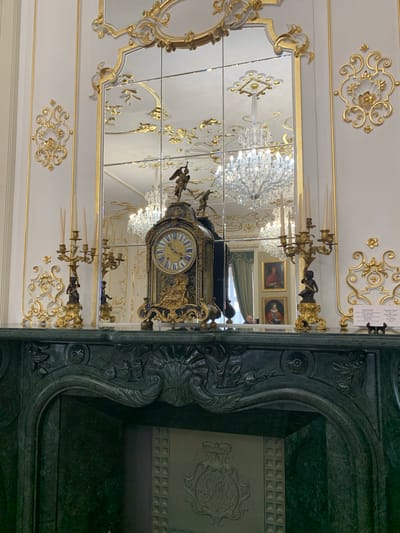 The image depicts an opulent interior within the Mir Castle Complex, located in Mirsky Sel'sky Sovet, Belarus. The central focus is a grand, dark green polished marble fireplace mantelpiece featuring intricate carved decorative elements, including shells, leaves, and floral motifs. The firebox opening is dark, revealing a light-colored interior panel adorned with a crowned "MR" monogram.
Resting on the mantelpiece are three ornate items. In the center is a large, dark-colored mantel clock with extensive gilded bronze ornamentation, including mythological figures and scrollwork. A golden winged figure stands atop the clock. The clock face is round, featuring blue Roman numerals on a white inner ring and a gold center. Flanking the clock are two identical gilded three-branched candelabras, each holding three upright white taper candles. The base of each candelabra features a dark statuette, possibly a putto or cherub. To the right of the right candelabra, a small informational sign written in Cyrillic rests on a miniature wooden easel.
Above the mantelpiece is a tall, segmented mirror composed of multiple rectangular panes, framed by a highly ornate, gilded border with elaborate Rococo-style scrollwork and floral designs. The mirror reflects a grand room beyond the immediate view, including multiple crystal chandeliers with numerous lit bulbs, indicating a high ceiling. Also visible in the reflection are two framed portrait paintings: an upper painting of a man in a red hat and a lower painting of a woman in formal attire and a crown. Green drapery is visible to the left of the reflected paintings. A partial reflection of an adult individual, identified by a dark shirt and a hand holding a mobile phone, is visible in the lower right segment of the mirror, suggesting a visitor capturing an image of the scene.
The surrounding walls are light-colored, possibly white or cream, adorned with vertical gilded decorative trim and ornate gilded appliqués featuring intricate floral and scroll patterns. The overall scene conveys a sense of historical grandeur and elaborate classical design.