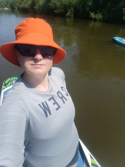 A woman is taking a selfie while on a paddleboard in a river. She is wearing an orange bucket hat and sunglasses, a grey long-sleeved t-shirt with "CREW" printed on it in dark letters, and dark shorts. A second paddleboard is visible in the water to the right of the frame. The background shows lush green trees lining the riverbank. This location is in the city of Berkuty, Russia.