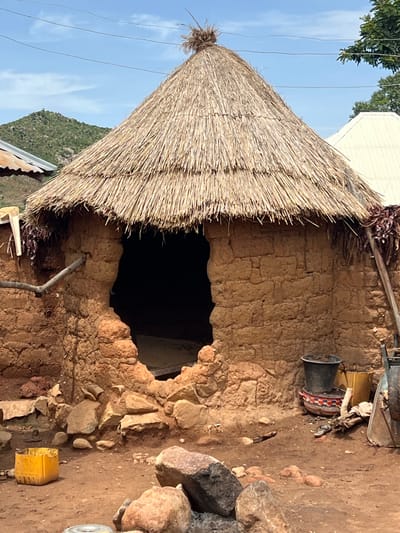 The image shows a traditional round hut with a thatched roof in Gwolong, Nigeria. The hut is constructed from mud bricks and has a large, irregular opening for an entrance. Rocks and dirt surround the base of the hut. In front of the hut, there are several objects, including a yellow plastic container, a black bucket, a colorful bowl, a pair of flip-flops, and a wheelbarrow. In the background, a green, hilly landscape with scattered trees is visible under a bright blue sky with some clouds.