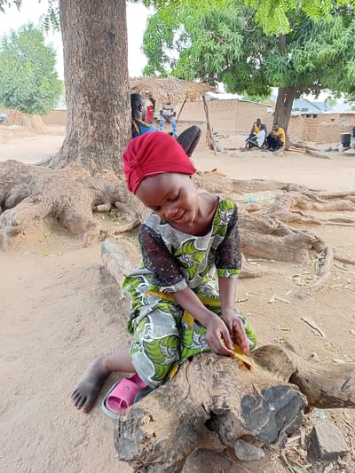 A young girl sits cross-legged on a weathered tree stump in Nata-Ala, Nigeria. She wears a vibrant green and white patterned dress with black lace sleeves and a red head covering. She holds a small yellow leaf in her hands. Her right foot is bare, while a pink sandal adorns her left. The stump is surrounded by sandy ground, and a large tree trunk extends upwards on the left side of the frame. In the background, a group of people gather near buildings under the shade of a tree.