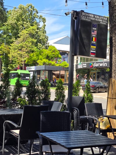The image shows an outdoor seating area with tables and chairs, likely part of a cafe or restaurant. In the background, a green bus is parked near a bus stop labeled "PANDA TOUR". A digital currency exchange board is prominently displayed, showing rates for EUR, USD, RON, UAH, and RUB. Next to the currency exchange is a building with a sign for "darwin" and "Beauty Shop". People are visible walking and sitting in the background. The scene is set on a sunny day with trees and a blue sky overhead. The architecture and signage suggest an urban environment in Eastern Europe, consistent with Edineț, Moldova.