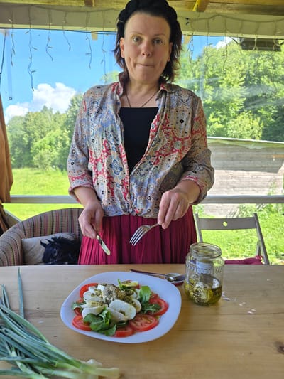 A woman stands behind a wooden table on a covered porch, holding a fork and knife, ready to eat a caprese salad. The salad is on a white plate and consists of sliced tomatoes, mozzarella balls, and arugula, drizzled with pesto and olive oil. Next to the salad is a glass jar containing marinated cheese balls in olive oil and a spoon. A bunch of green onions lies on the table to the left of the plate. In the background, through a large window, a lush green landscape with trees and a clear blue sky with white clouds is visible. A patterned armchair with a cushion depicting a black horse is partially visible to the left. The woman is wearing a black headband, a black t-shirt, and a colorful, patterned open shirt over it, paired with a dark red skirt. The location is identified as Никитское, Russia.